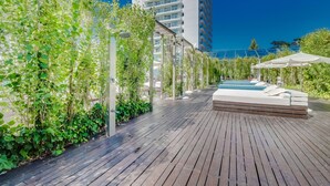 Une terrasse en bois avec une piscine, entourée de verdure et avec un grand immeuble en arrière-plan.