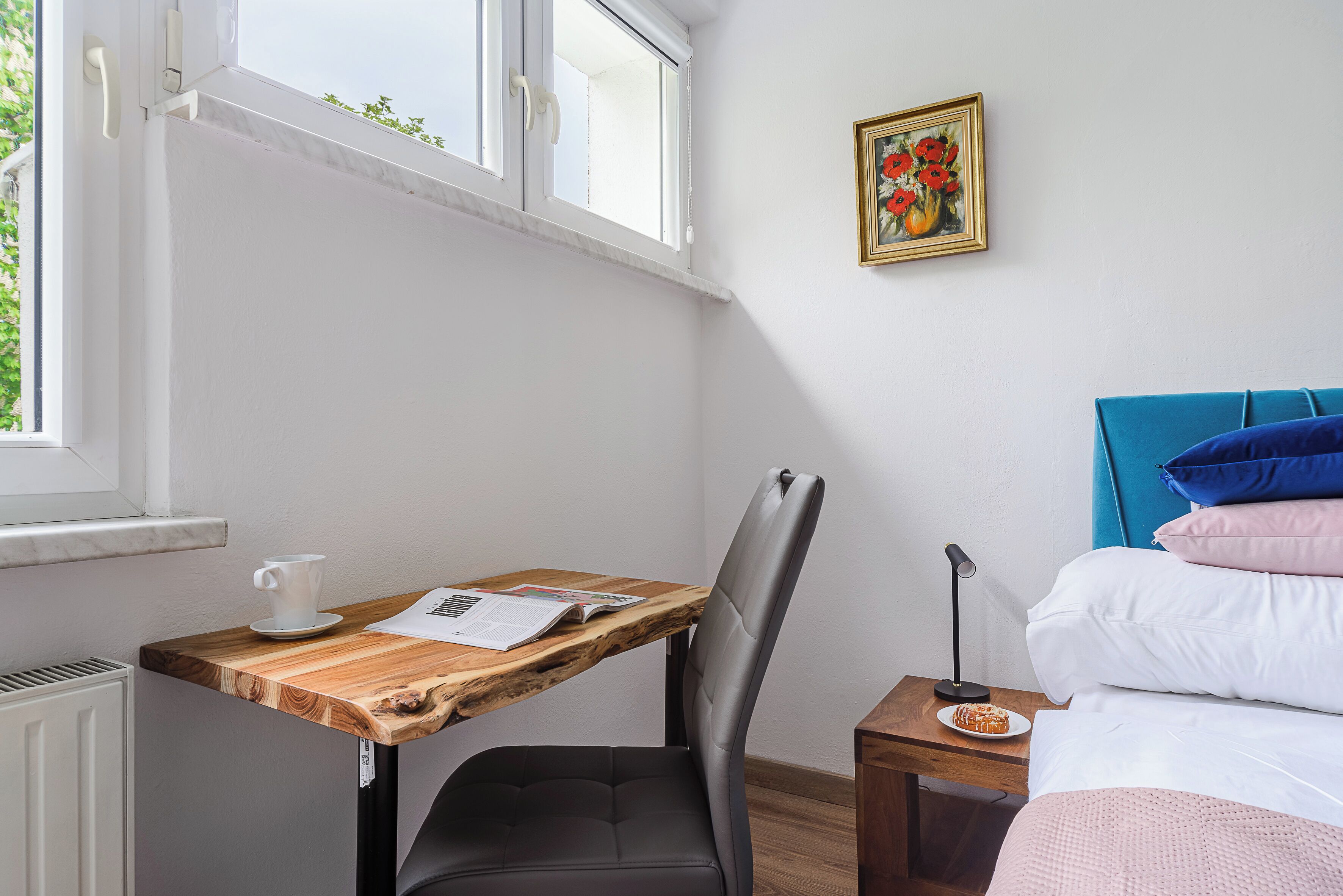 Una habitación con un escritorio de madera, una silla, una cama con almohadas azules y rosas, un cuadro enmarcado y una ventana con vistas a zonas verdes.