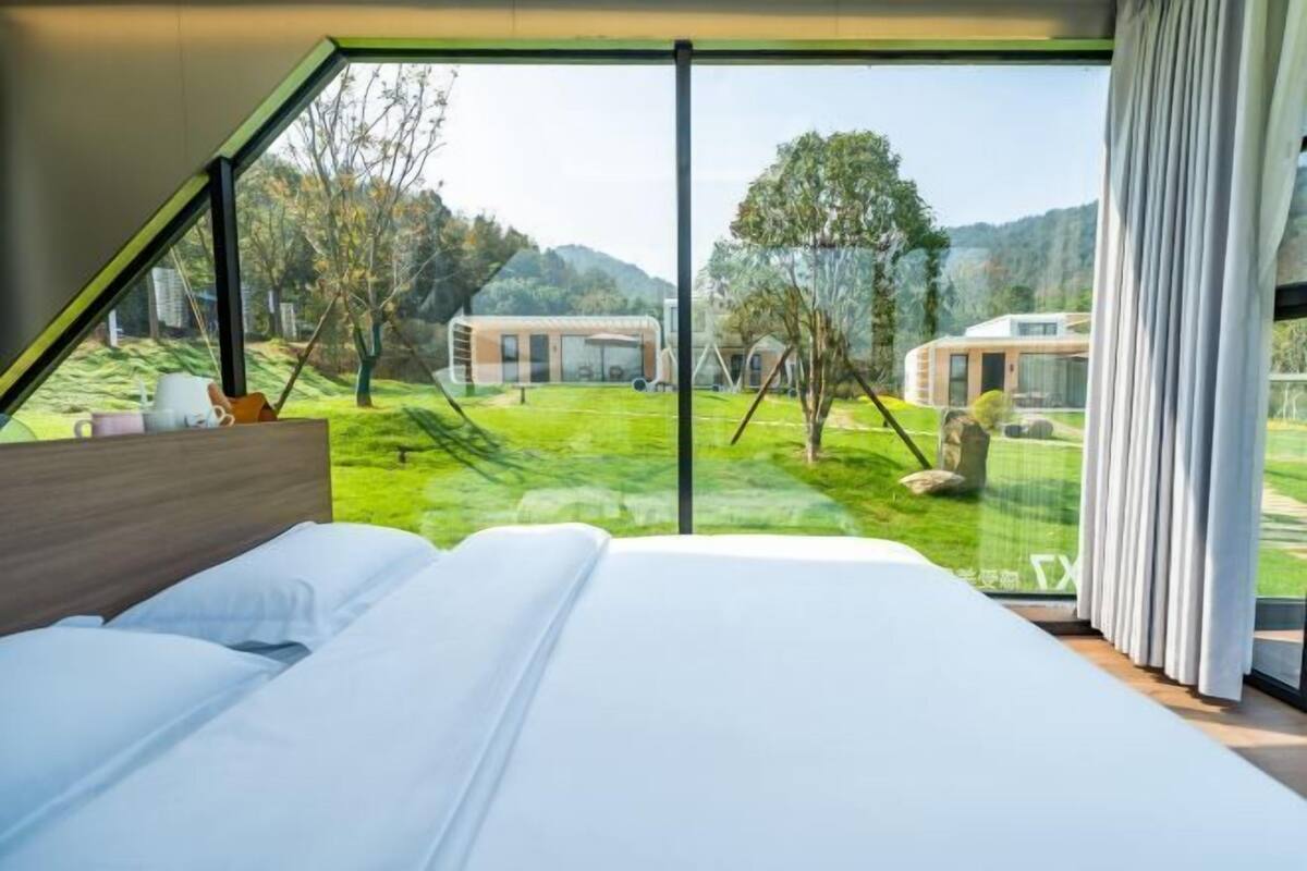 Une chambre avec une grande fenêtre, un lit et une vue sur un paysage verdoyant et des bâtiments.