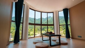 Een kamer met grote ramen en uitzicht op een bergachtig landschap, een klein tafeltje en rondzitgedeeltes.