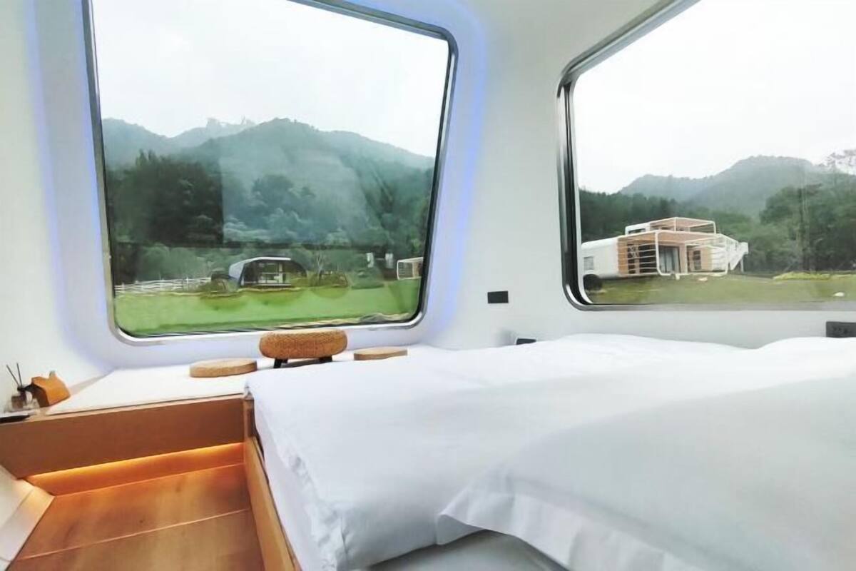 Une chambre moderne avec un lit, de grandes fenêtres et une vue sur une maison et des montagnes.