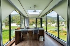 Een moderne keuken met een groot eiland, barkrukken en uitzicht op een groen landschap door grote ramen.