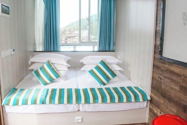 A single bed with white bedding and striped pillows, a window with blue curtains, and a red bucket on the floor.