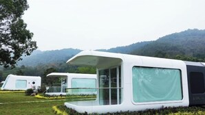 A series of modern, white, capsule-style accommodations with large windows, set in a grassy area with a mountainous backdrop.