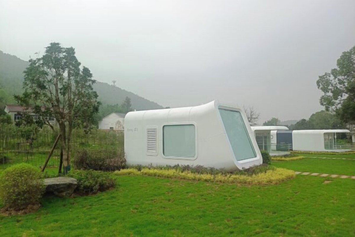 A modern, white, curved structure with a window, set in a grassy area with trees and a hill in the background.