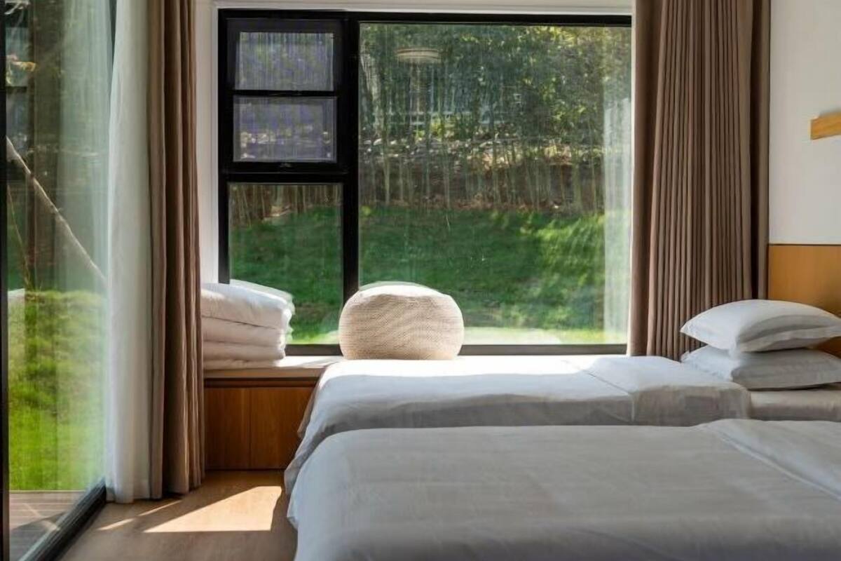 Une chambre avec une grande fenêtre, un lit, une table de chevet et une vue sur la verdure.