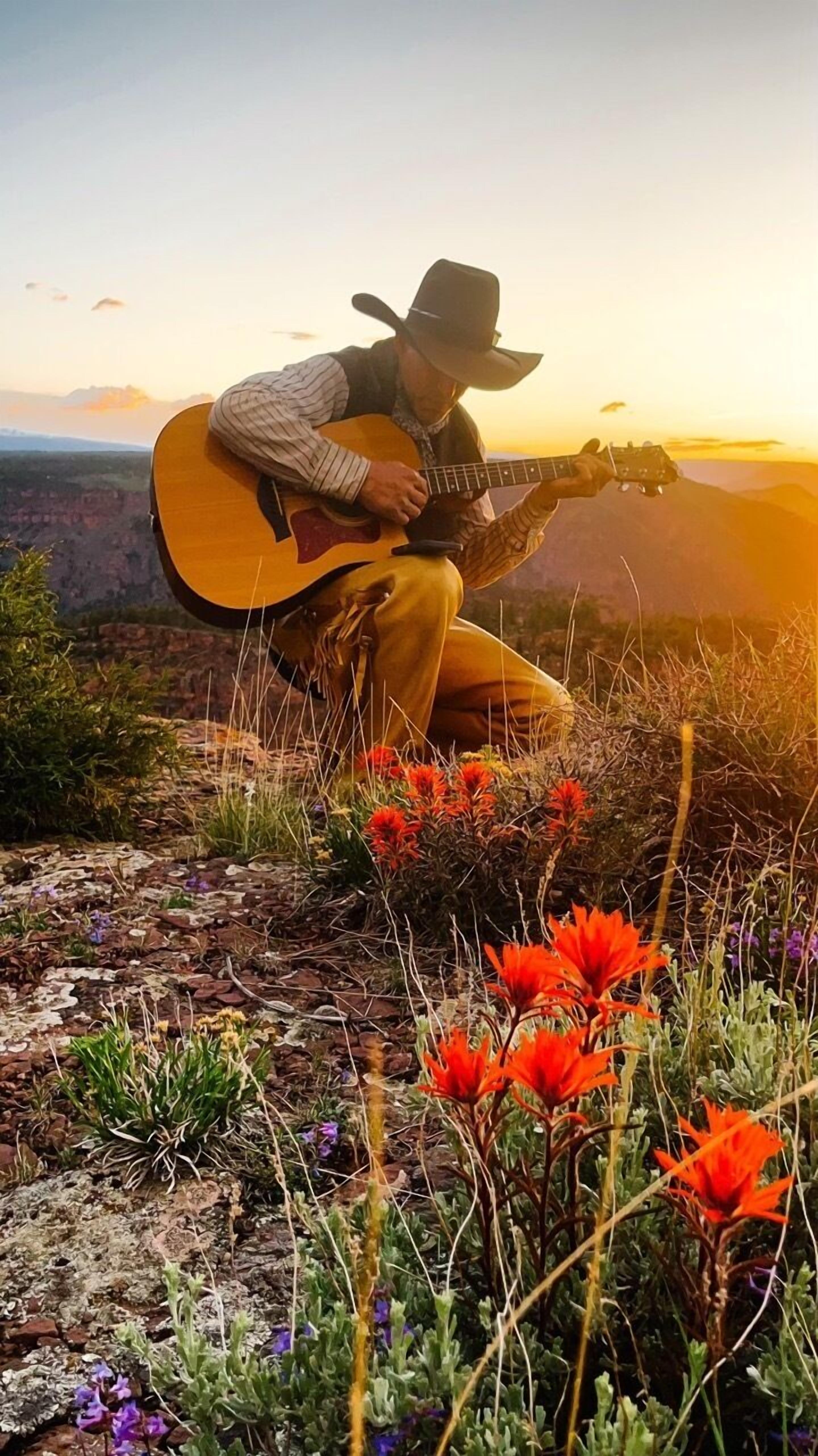 Una persona tocando la guitarra sobre terreno rocoso, con flores y un atardecer de fondo.