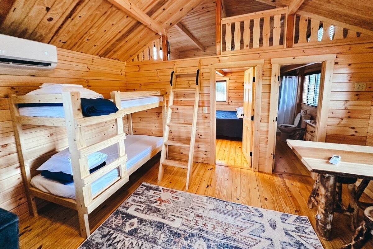 A wooden cabin interior with bunk beds, a dining table, and a visible bathroom.
