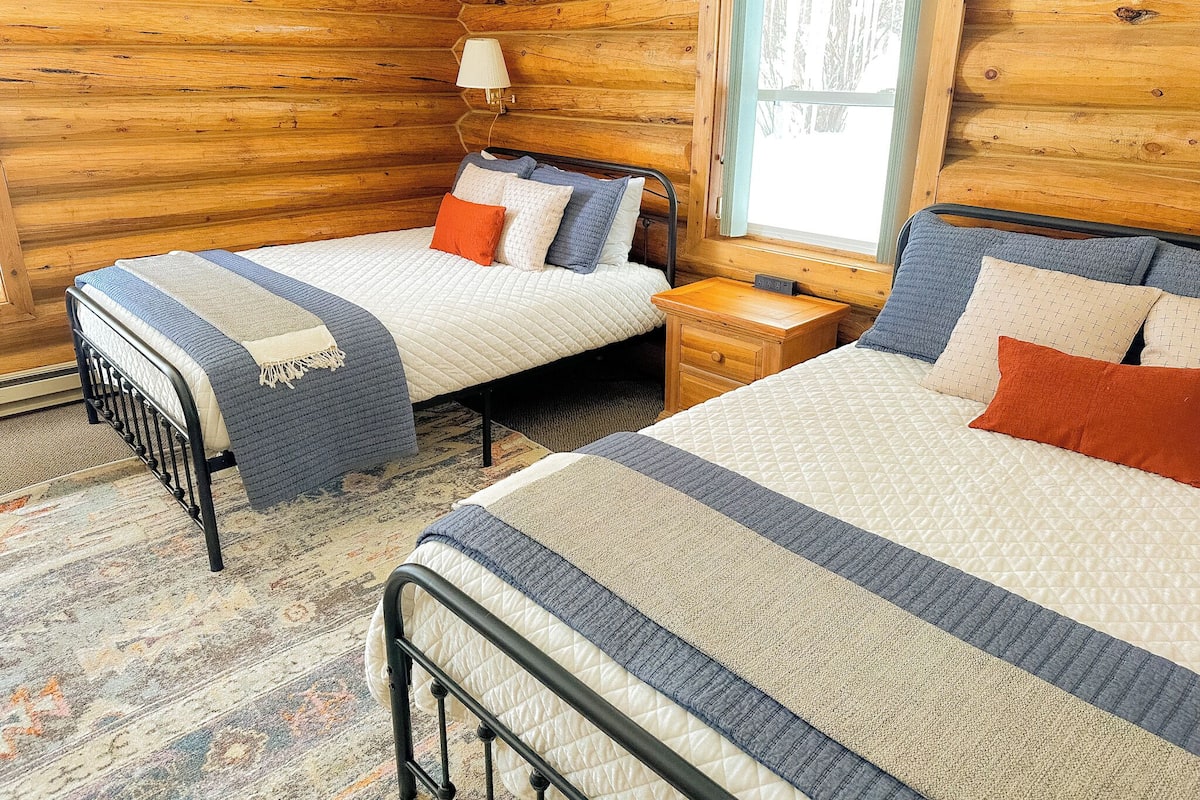 A cabin-style room with two beds, wooden walls, a window, and a nightstand with a lamp.