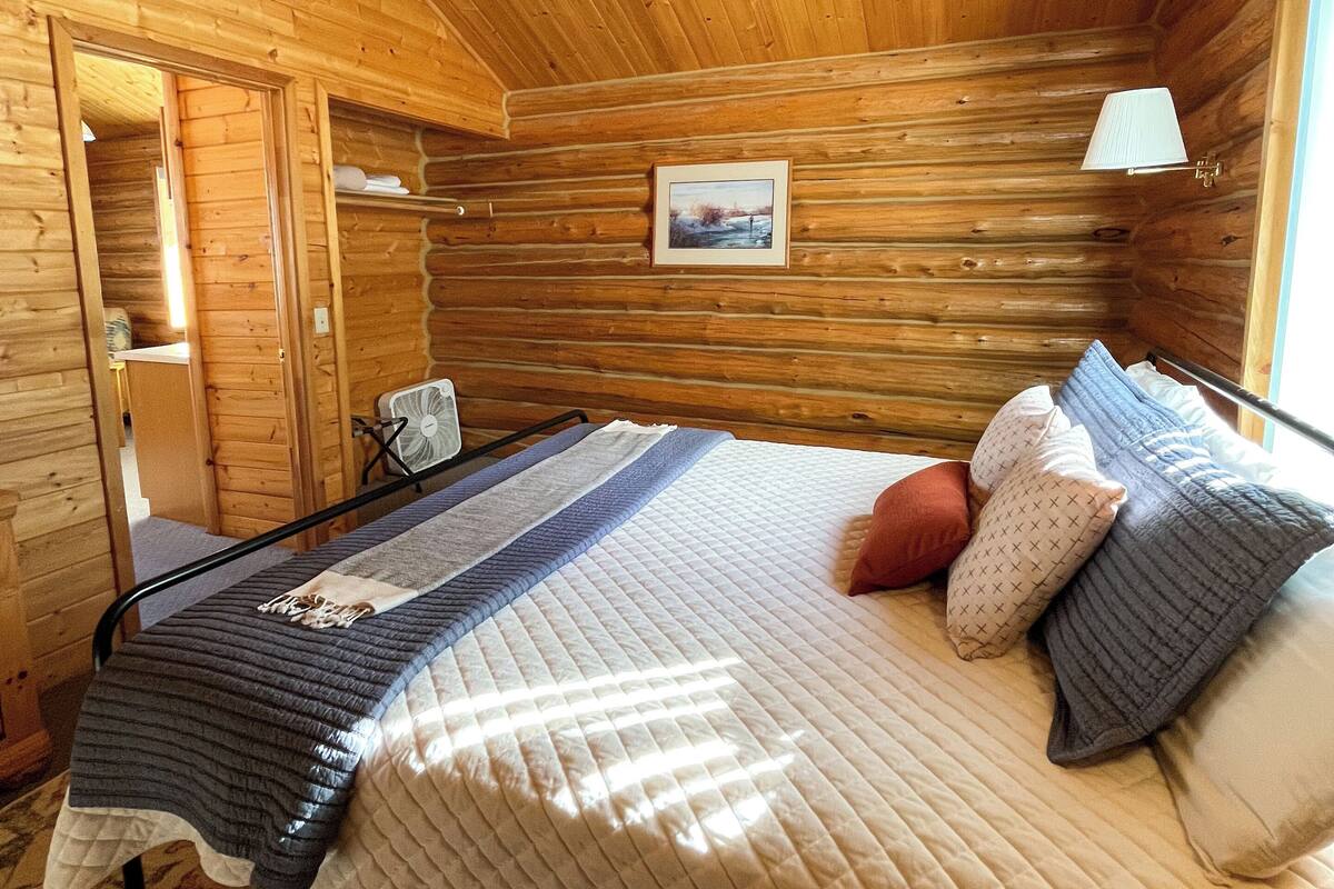 A wooden cabin room with a bed, a fan, and a framed picture on the wall.