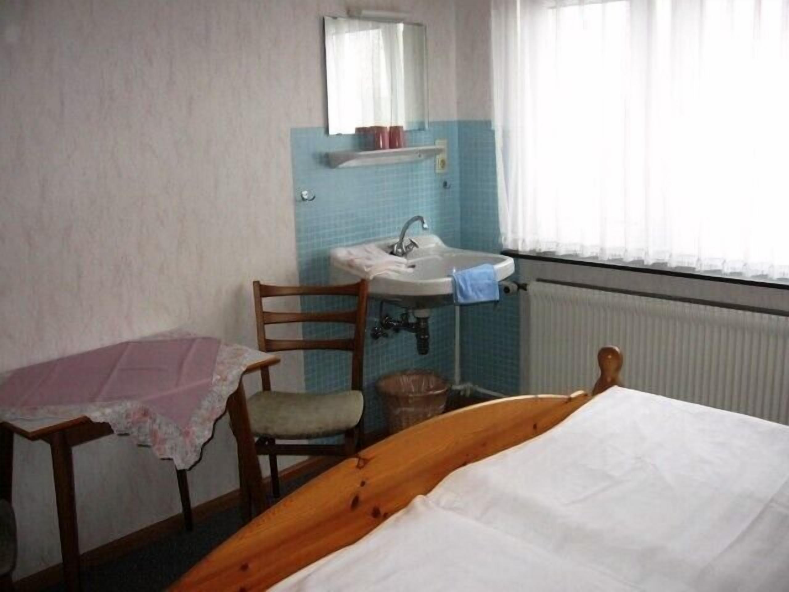 Ein Schlafzimmer mit einem hölzernen Bett, einem kleinen Tisch mit rosa Tischdecke, einem Stuhl, einem Waschbecken und einem Fenster mit Vorhängen.