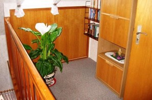Een hal met een houten trap, een potplant, een boekenplank en een deur met de tekst 