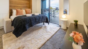 Ein modernes Schlafzimmer mit einem großen Bett, einem kleinen Nachttisch, einem bei dem Bett stehenden Tisch, einem großen Fenster und einem Balkon.