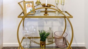 Ein goldfarbener, zweistöckiger Wagen mit Glasbögen, der eine Kerze, Champagnergläser, einen Bilderrahmen, eine kleine Pflanze und Glaswaren präsentiert.