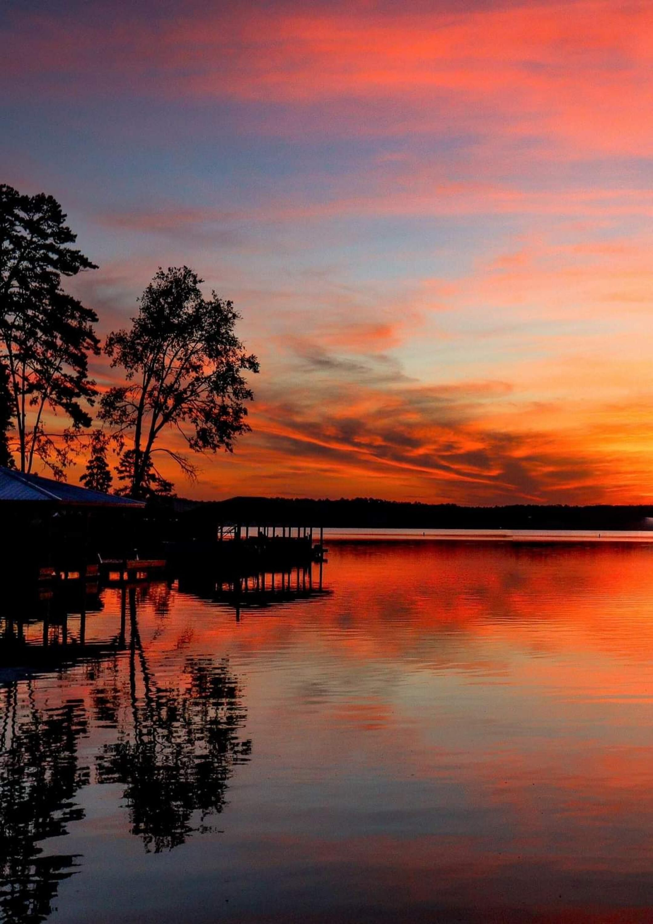 A calm lake at sunset with a pier and trees on the left.