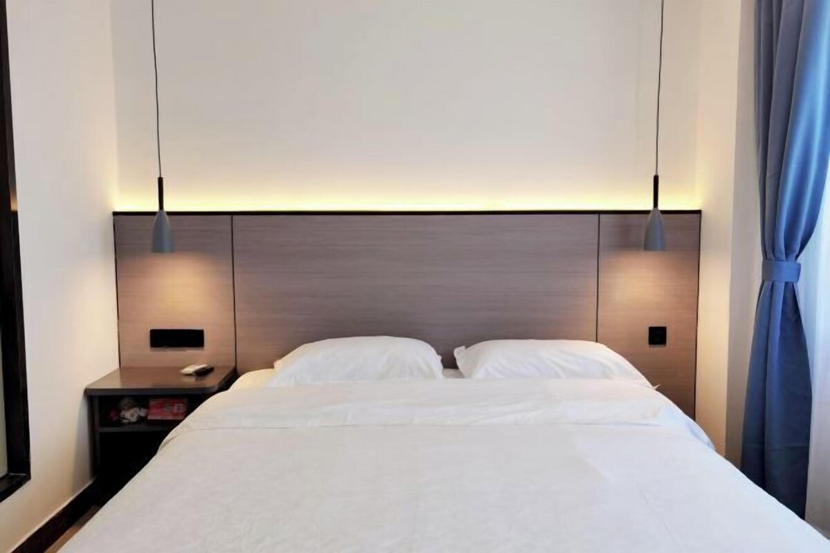 A modern hotel room with a neatly made bed, a bedside table with a lamp, and a headboard with integrated lighting.
