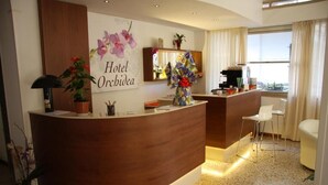 A hotel reception area with a wooden reception desk, a large floral artwork, and a view of the city through the window. - Hotel Orchidea (Rimini)