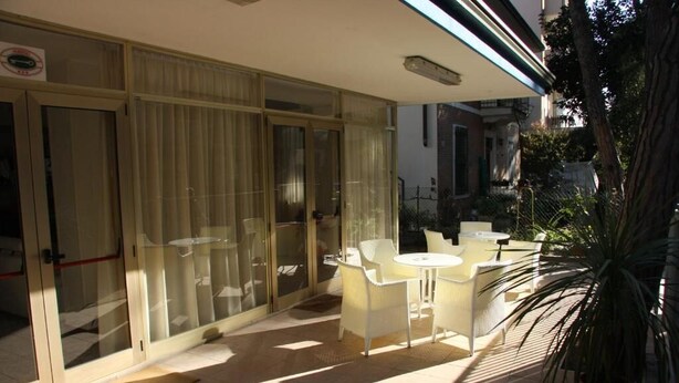 A sunlit patio area with a table and chairs, a potted plant, and a glass door leading to a room. - Hotel Orchidea (Rimini)