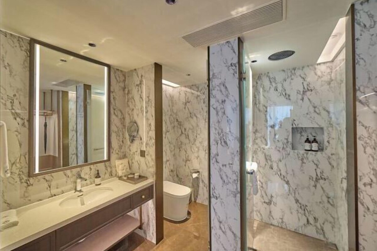 Un baño con pared de mármol, un espejo grande, un lavamanos y un inodoro.