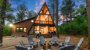 Una cabaña con techo triangular, una terraza y una fogata rodeada de sillas Adirondack.