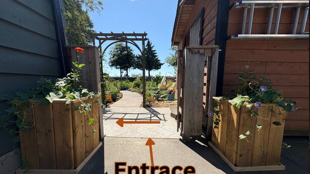 Une porte d’entrée en bois, avec un chemin menant à un jardin, le tout entouré de clôtures en bois et de plantes.