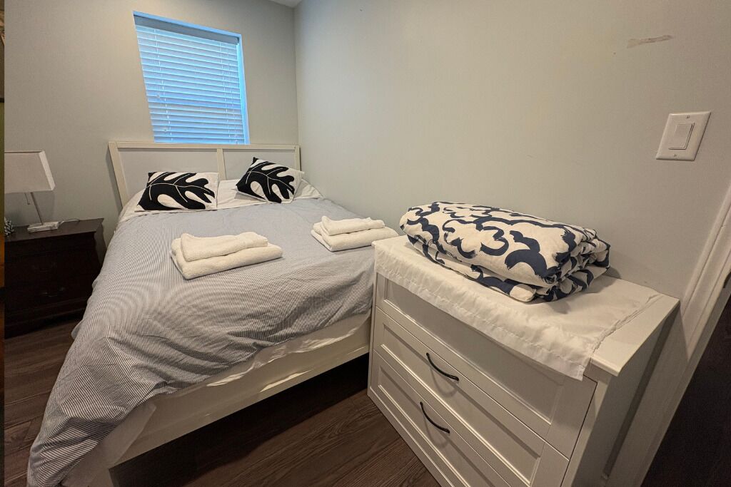 Cama bem arrumada com manta listrada e duas almofadas decorativas, mesa de cabeceira com abajur, e janela com persianas.