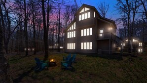 A large, multi-story house with a prominent balcony, surrounded by trees and a grassy area with two chairs and a fire pit.