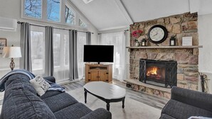 A living room with a stone fireplace, a flat-screen TV, and a large window with curtains.