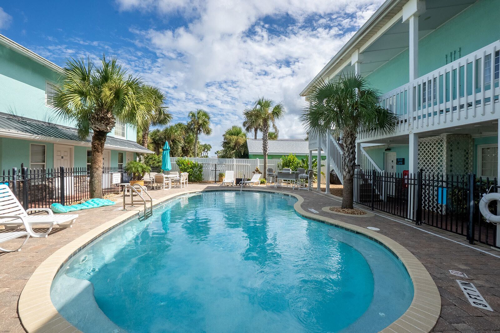 A swimming pool with lounge chairs and umbrellas, surrounded by palm trees and a building with a white fence.