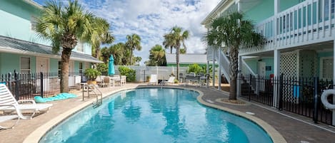A swimming pool with lounge chairs and umbrellas, surrounded by palm trees and a building with a white fence.