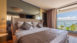 Quarto de hotel moderno com uma cama grande, mesa de cabeceira, espelho e vista para o mar.