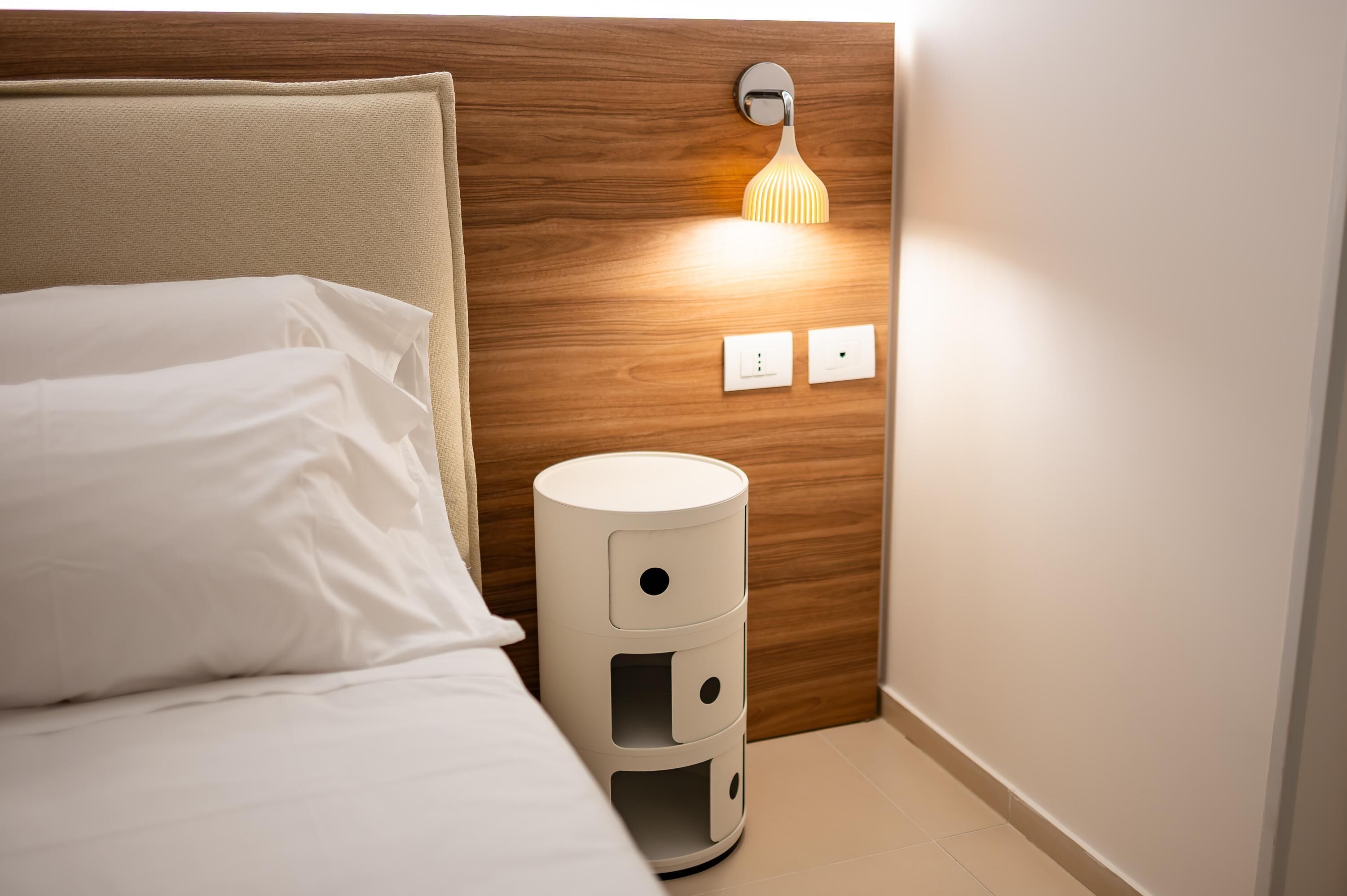 Un lit bien fait, avec du linge de lit blanc, une lampe de chevet et une prise de courant fixée au mur.