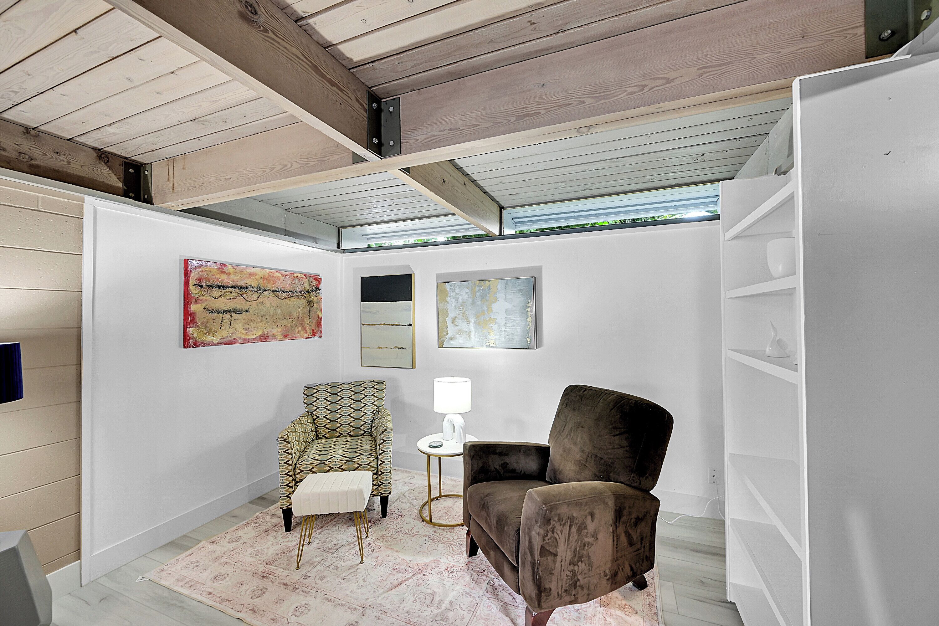 A room with a wooden ceiling, a white wall with artwork, a patterned armchair, a dark armchair, a small white table with a lamp, and a white shelving unit.