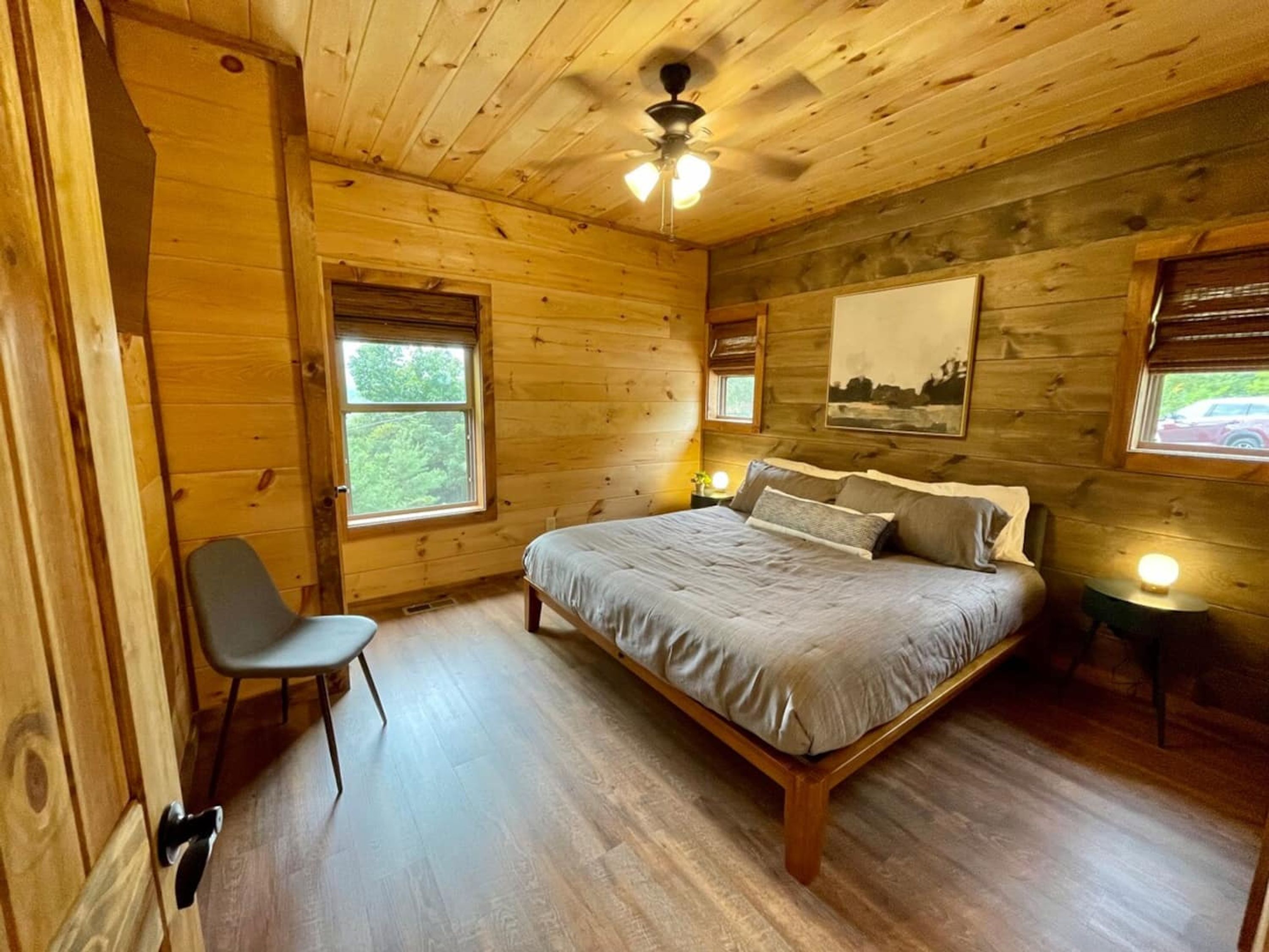 A wooden cabin bedroom with a bed, a chair, and two windows.