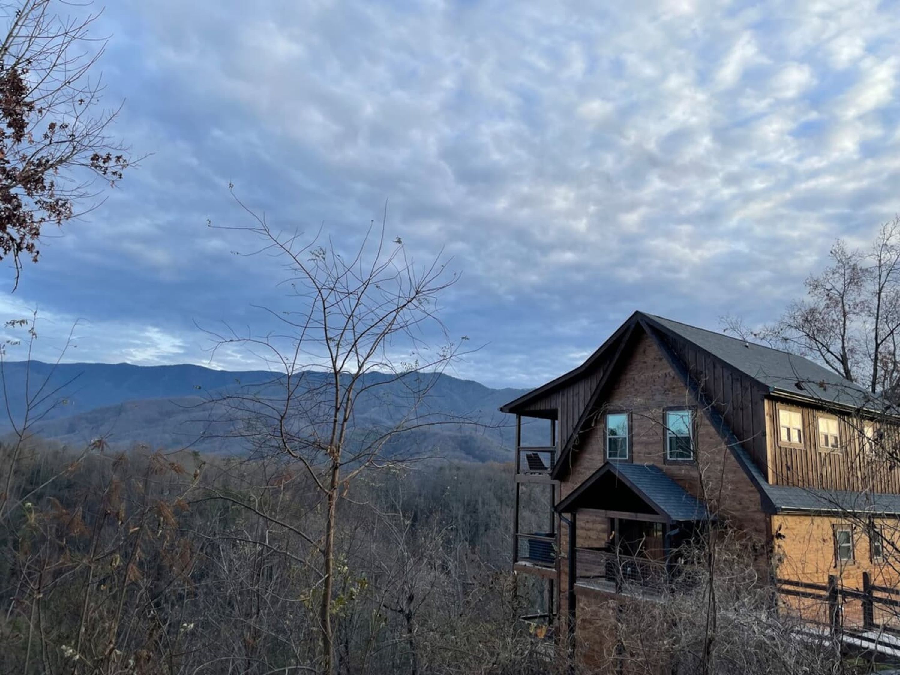 A two-story wooden cabin with a balcony, surrounded by a forest and mountains in the background.