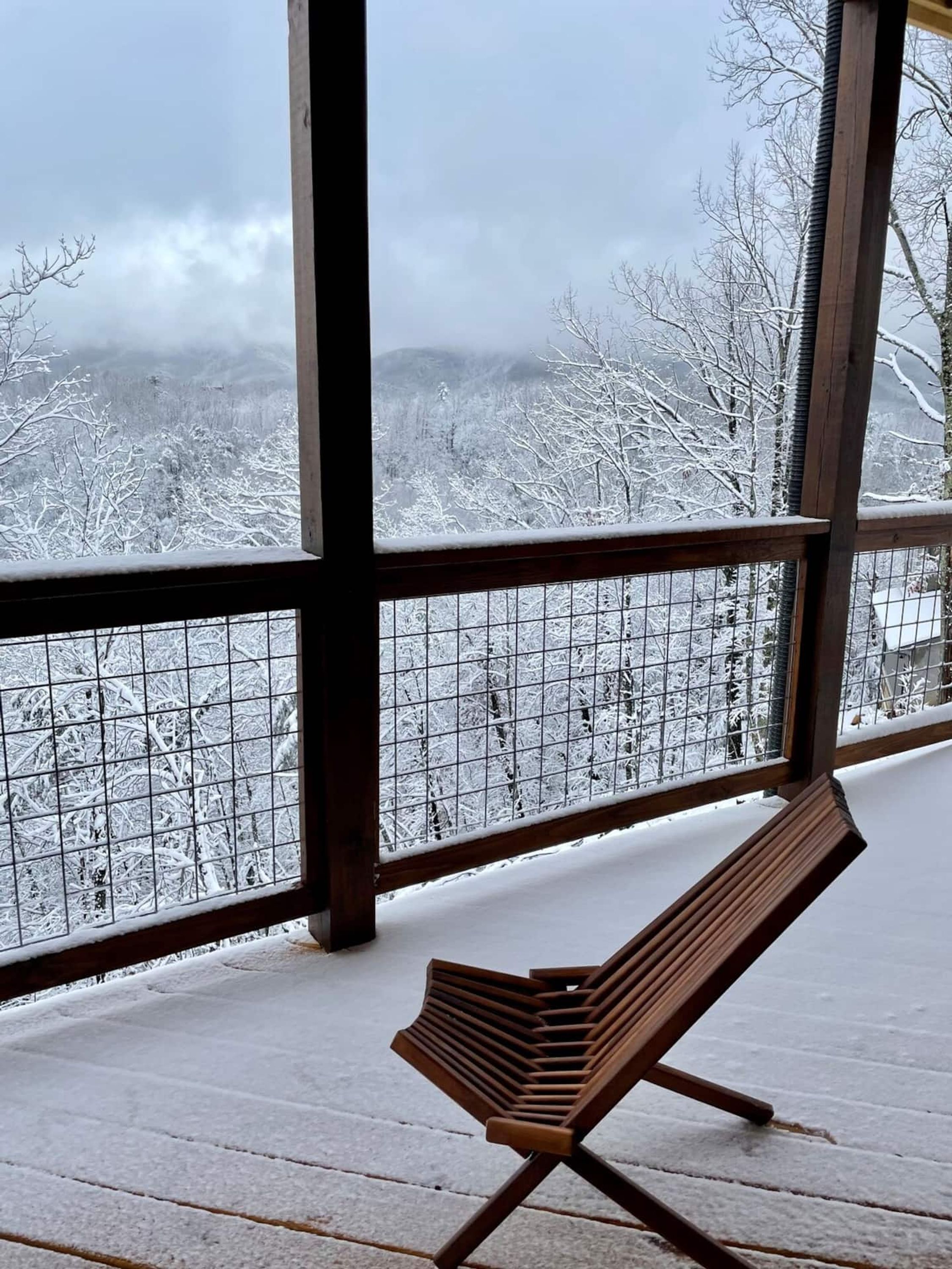 A wooden bench on a snow-covered deck with a view of snow-covered trees and mountains.