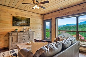 Ein gemütliches Wohnzimmer mit Holzbalkendecke, einem großen Fenster mit Blick auf die Berge, einem Ledersofa und einer Holzkabine mit Dekorationen.