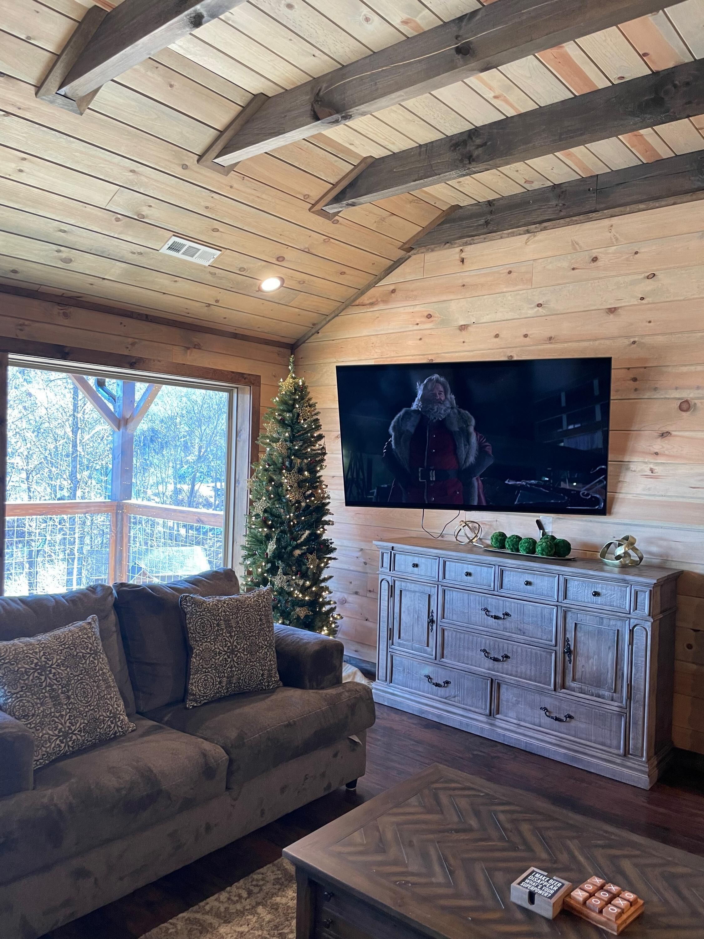 Una sala de estar acogedora con techo de madera, un televisor de pantalla plana grande, un árbol de Navidad y un sofá con cojines.