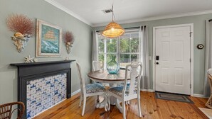 A dining area with a round table, white chairs, a hanging light fixture, a fireplace with a tiled backsplash, and a door with a doormat. - Frog Cottage (Beaufort)