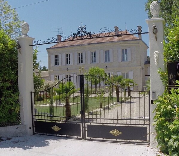 A gated entrance to a building with a symmetrical facade, a tiled roof, and a well-maintained garden with palm trees. - Le Manoir Lacustre Medoc, large house near castles and Ocean15 to 22 people (Queyrac)