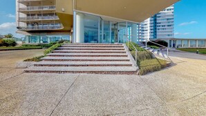 Un edificio moderno con fachada de vidrio, una serie de escalones de piedra que conducen a una entrada cubierta, y un cielo despejado.