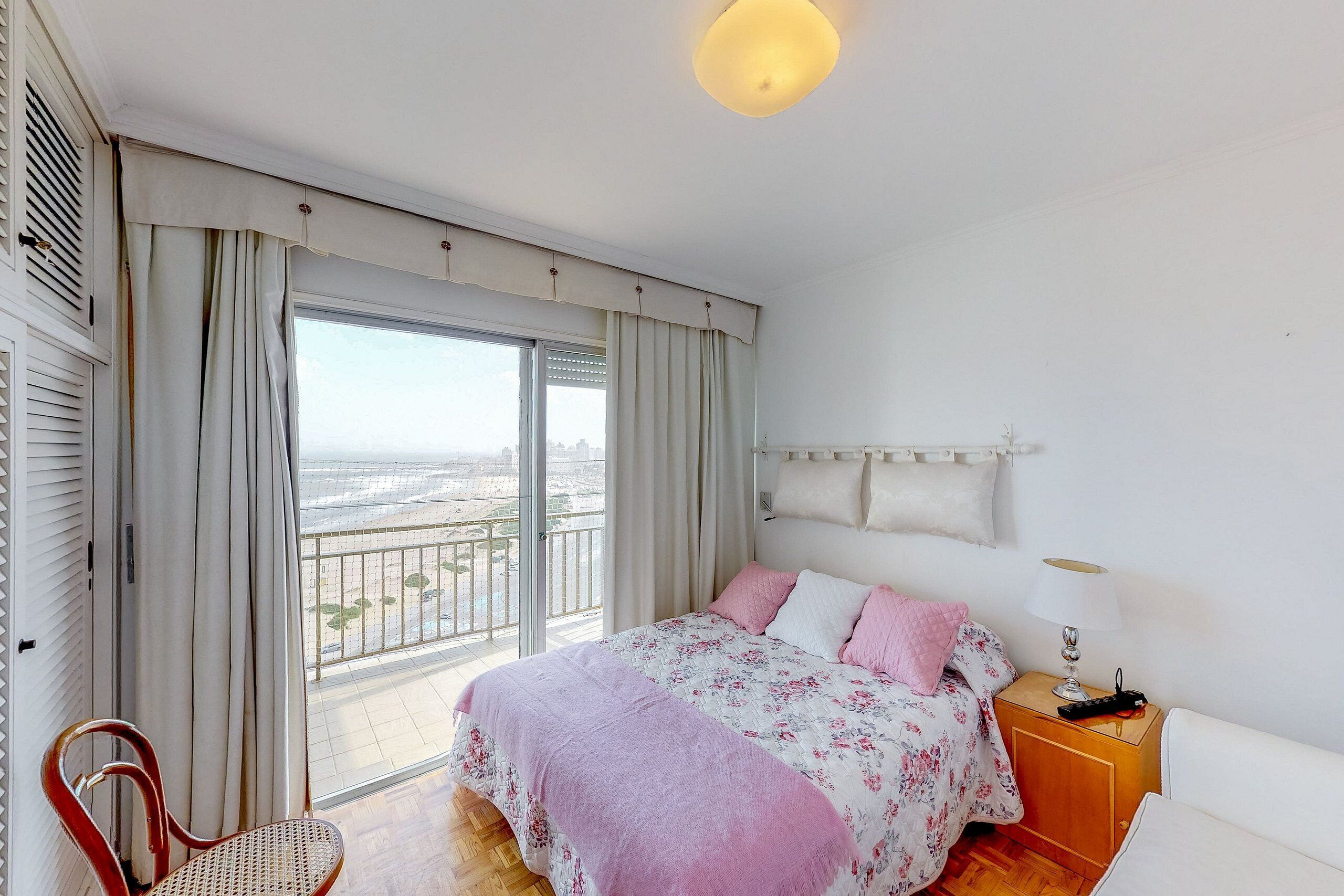 Ein Schlafzimmer mit Bett, Sessel, Nachttisch, Lampe und Blick auf die Stadt durch ein Fenster.