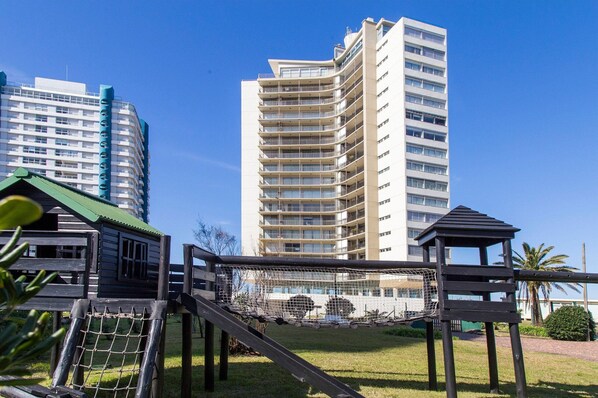 Un parque infantil con un tobogán y estructuras para escalar, rodeado de césped y un edificio alto al fondo.