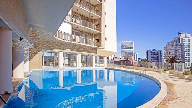 Una piscina con agua cristalina azul, rodeada por un edificio con balcones y un paisaje urbano al fondo.