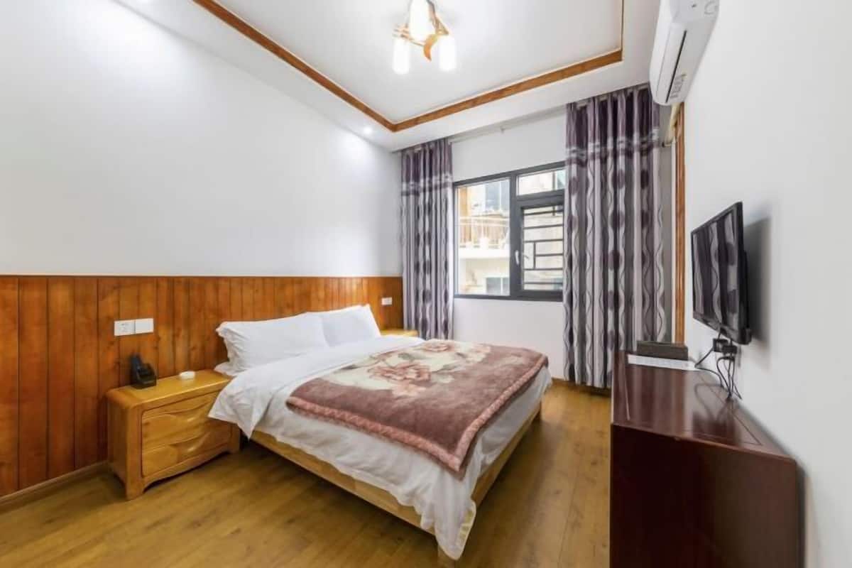 A bedroom with a wooden headboard, a bed with a floral bedspread, a nightstand, a TV on a stand, and a window with curtains.