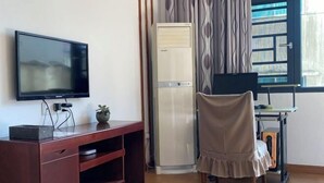 Quarto com televisão em suporte de madeira, cadeira bege, ar condicionado branco e janela com cortinas estampadas.