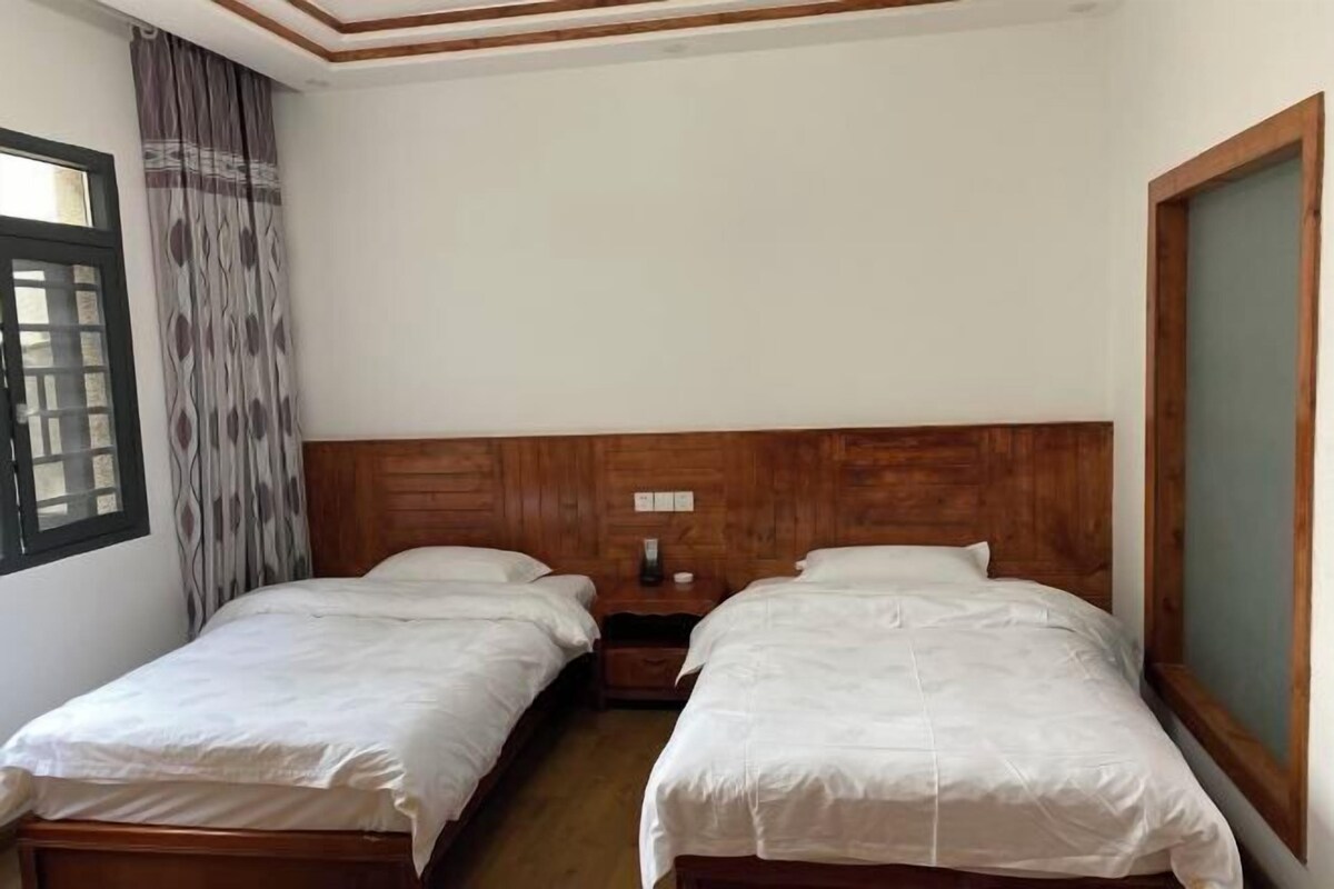 A room with two single beds, wooden headboards, a nightstand with a lamp, and a window with patterned curtains.