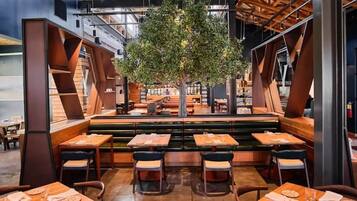 A modern restaurant interior with high ceilings, exposed beams, and a central tree. Tables are arranged around the seating area, with a mix of booths and standard tables.