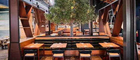 Un interior de restaurante moderno con techos altos, vigas a la vista y un árbol central. Mesas dispuestas alrededor del área de estar, con una mezcla de cabinas y mesas estándar.