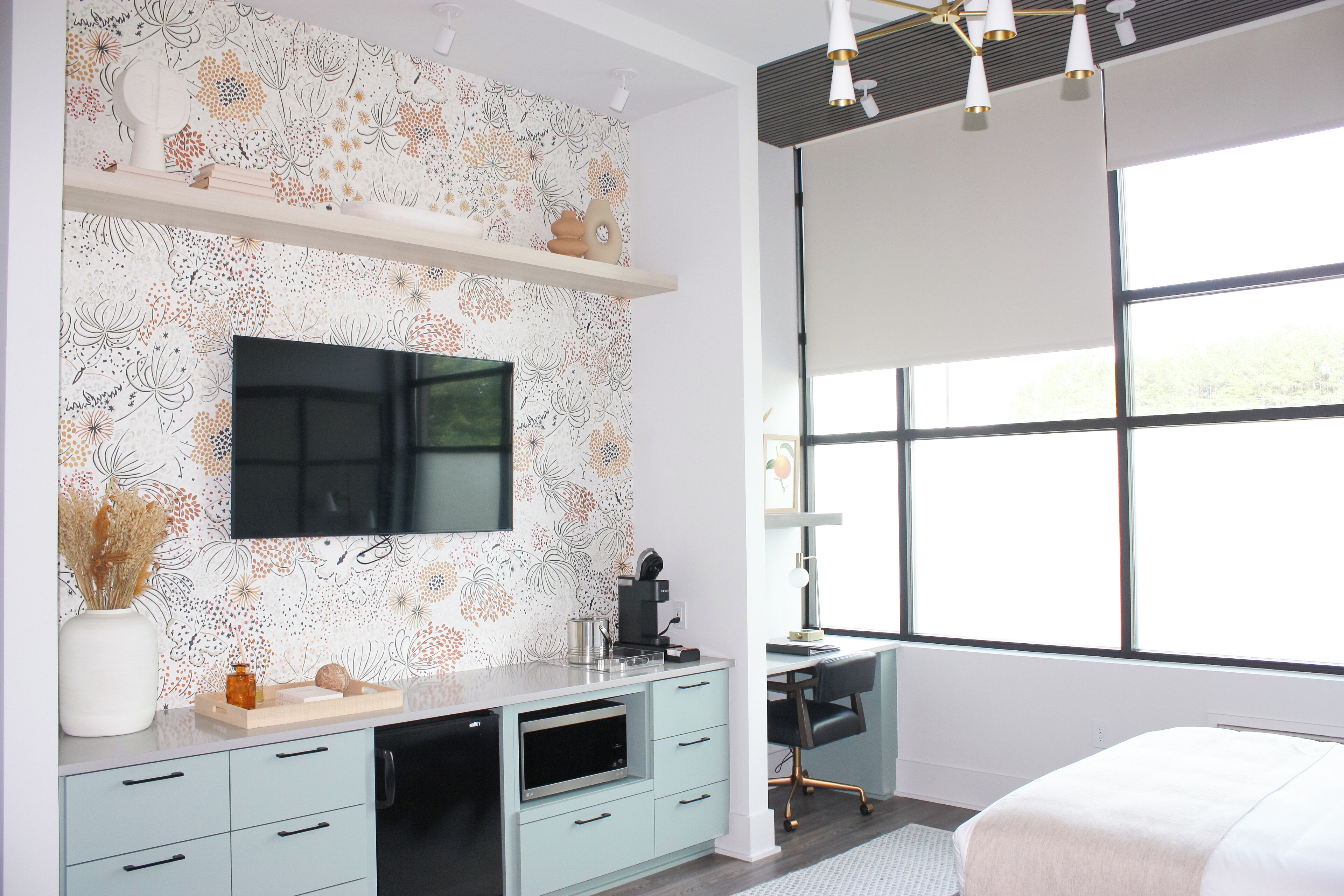 A modern bedroom with a large window, a flat-screen TV mounted on the wall, a desk with a chair, and a bed.